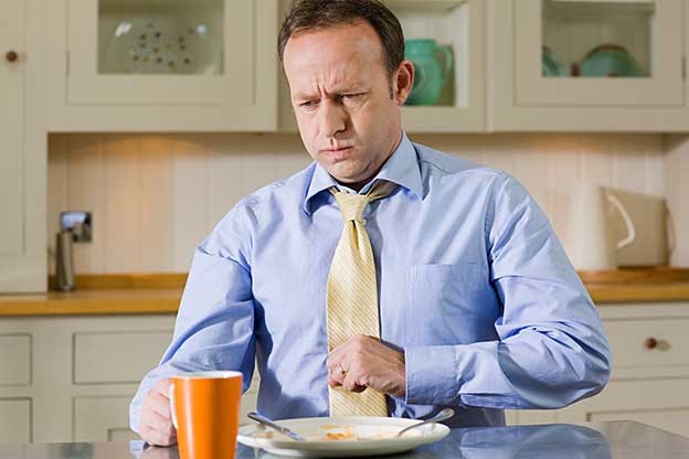 Man sitting at table in front of plate and glass, holding his stomach as if he has bloating, heart burn or indigestion.