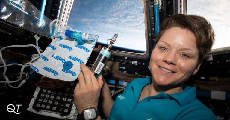 An astronaut holds a Quintron breath testing kit is pictured in space, with a glimpse of the Earth in the background.