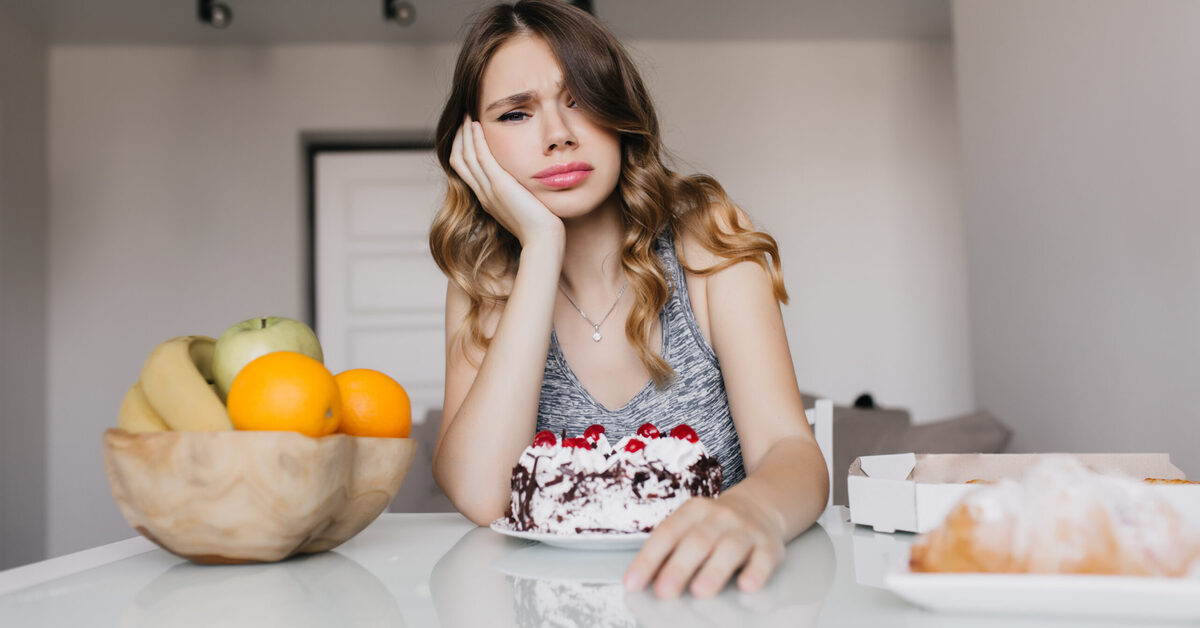 Young lady sitting at table with cakes and fruits contemplating which to eat.