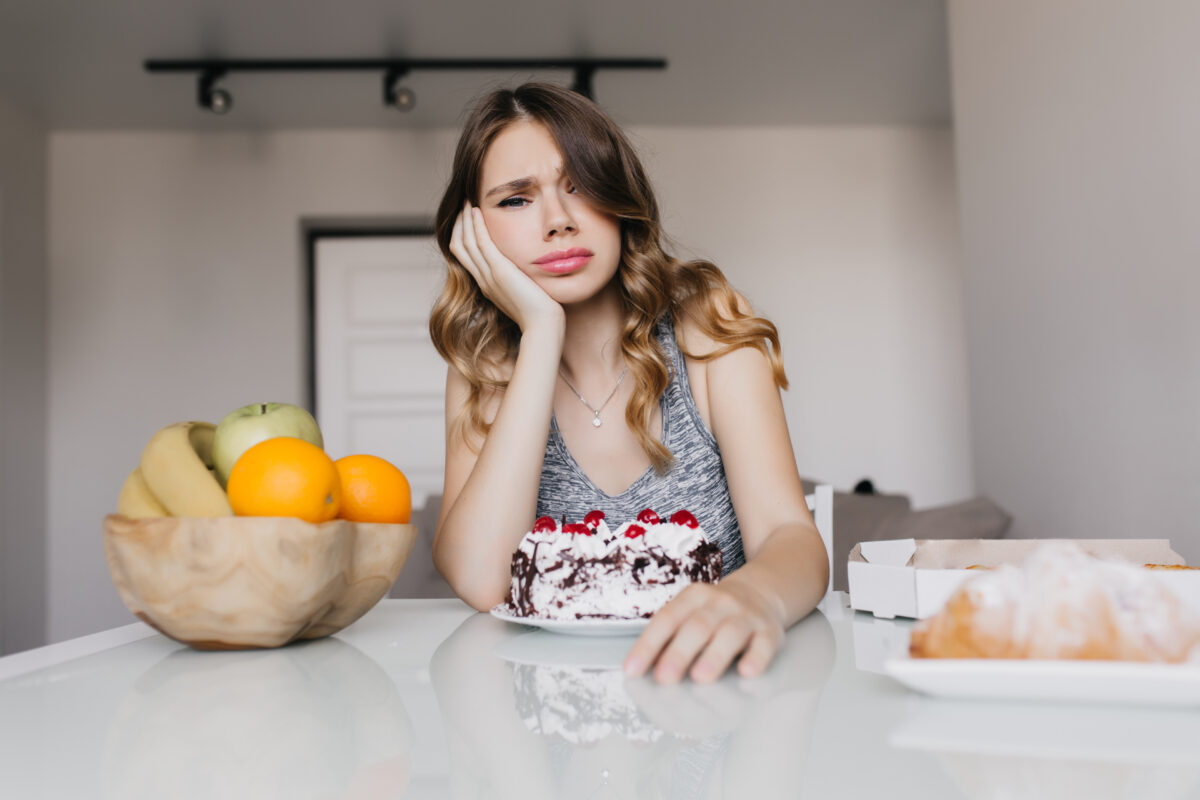 Young lady sitting at table with cakes and fruits contemplating which to eat.