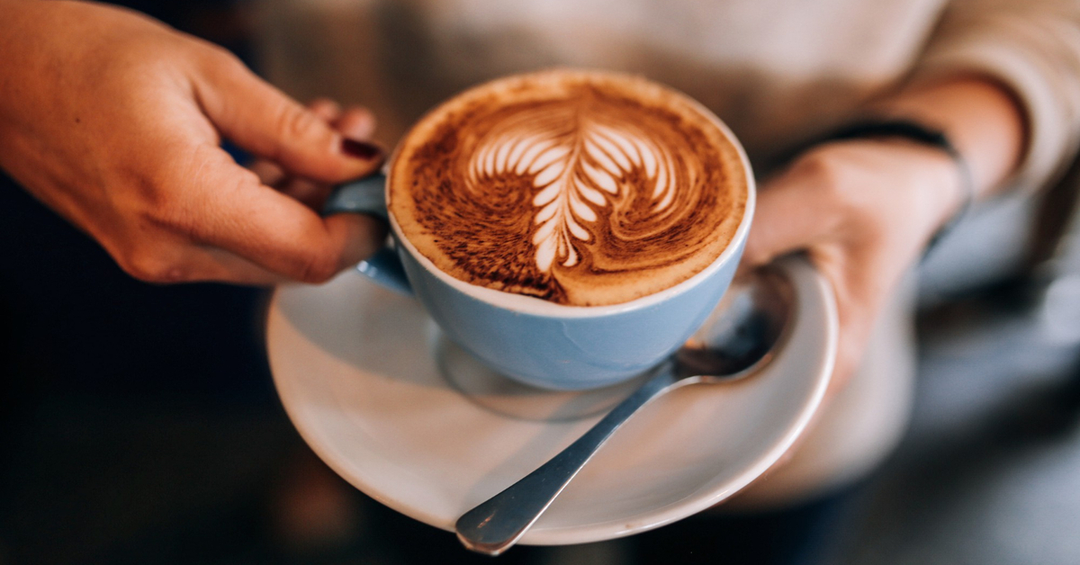 A person holds a cup of coffee on a saucer. The coffee is topped with steamed milk foam latte art.
